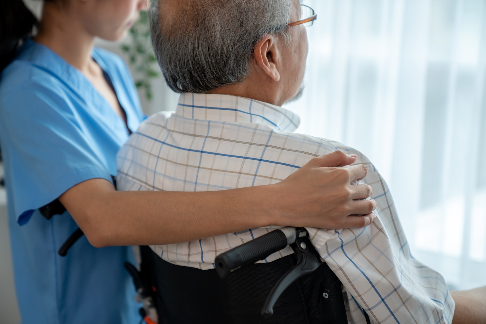 Caregiver resting a hand on an older Asian man's shoulder beside a bright window