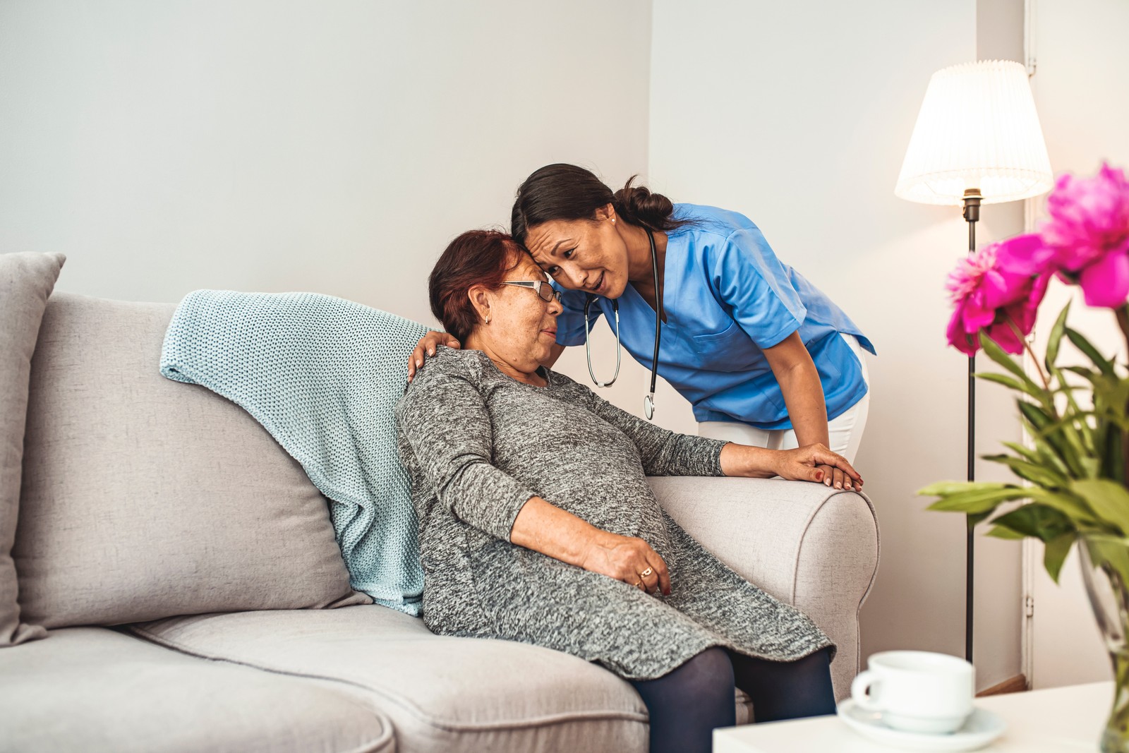 Nurse in blue scrubs embracing an older woman seated on a light sofa at home