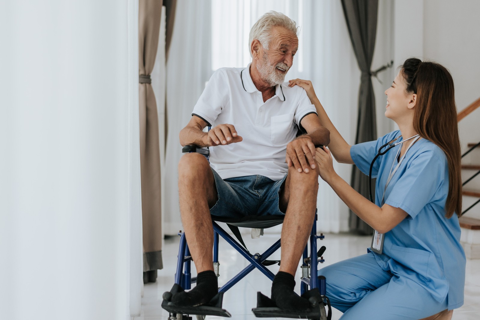 Physiotherapist checking an older man's knee while he sits in a wheelchair in a bright home